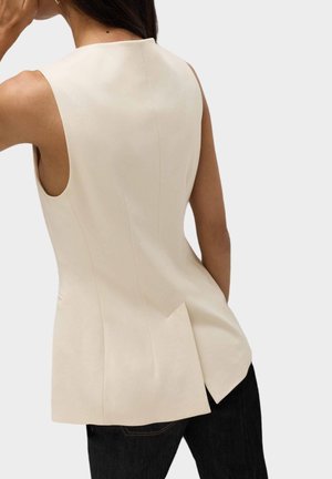 Haut sans manches crème au design ajusté, doté d'une pliure à l'arrière et d'une texture lisse. Associé à un jean foncé.