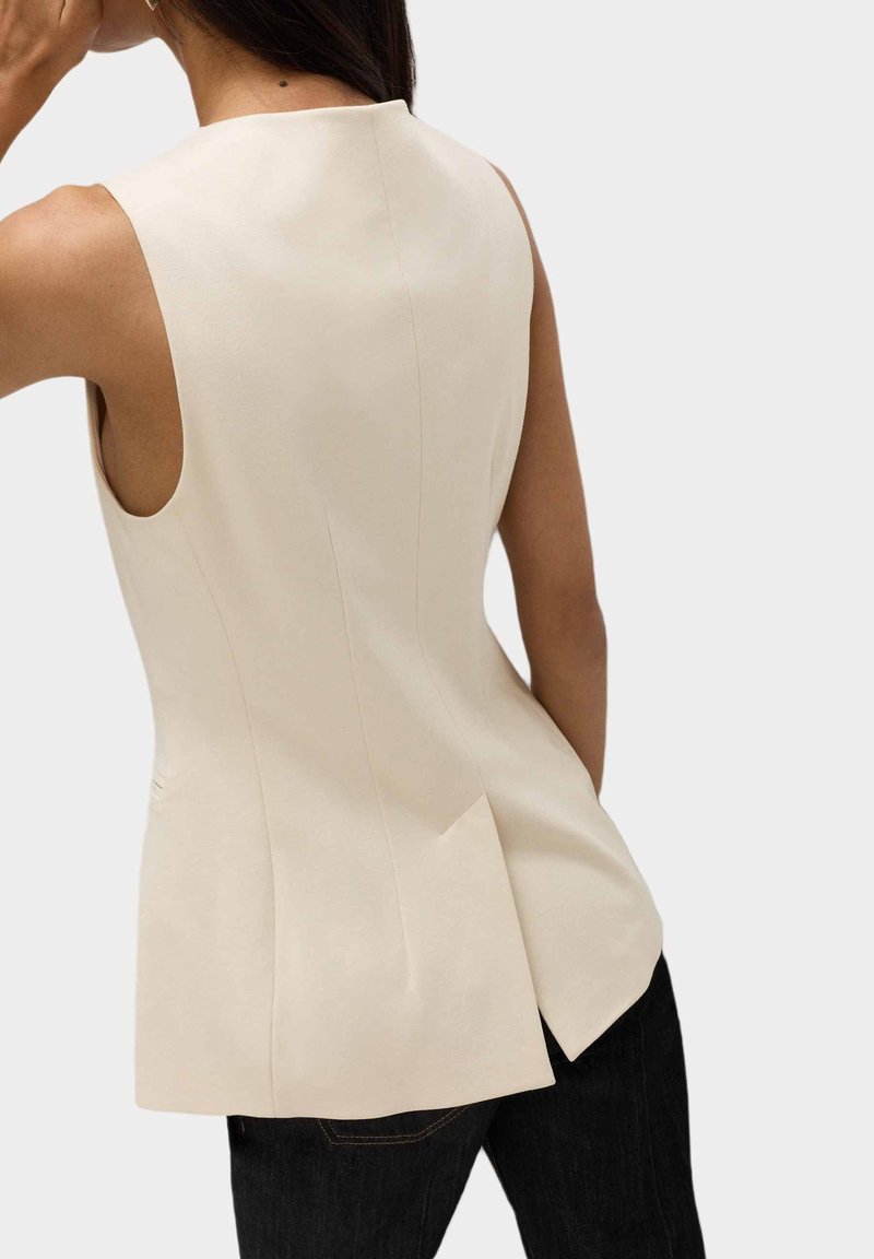 Creme ærmeløs top med et tætsiddende design, der har en plisse ved ryggen og en glat tekstur. Samlet med mørke jeans.