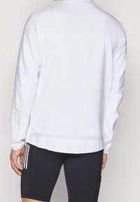 Veste de sport blanche à manches longues avec un col montant, une texture lisse, de subtiles perforations et un ourlet droit, associée à un short noir.