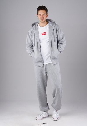 Graue Zip-Hoodie mit frontalen Taschen, kombiniert mit passenden grauen Jogginghosen. Das Modell trägt ein weißes T-Shirt mit einem roten Logo. Weiße Sneakers.