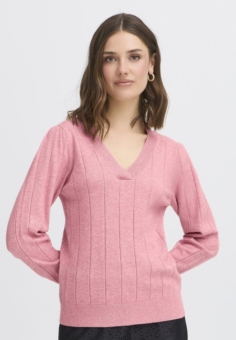 Vállig érő barna hajú nő arany karika fülbevalót visel és egy rózsaszín, V nyakú bordás pulóvert, amely egy egyszínű világos háttér előtt áll.