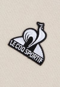 Logo ricamato in bianco e nero con un uccello stilizzato su uno sfondo beige chiaro, con "LE COQ SPORTIF" in lettere grasse.