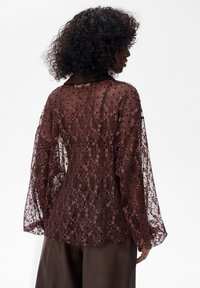 Przezroczysta burgundowa bluzka z misternym wzorem koronkowym, długimi rękawami i kwiatowymi motywami. Lekka tkanina ukazuje delikatną fakturę i akcenty.