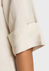 Chemise en lin avec manches retroussées, de couleur beige doux. Dispose d'une petite étiquette métallique de la marque cousue dans le poignet. Texture lisse.