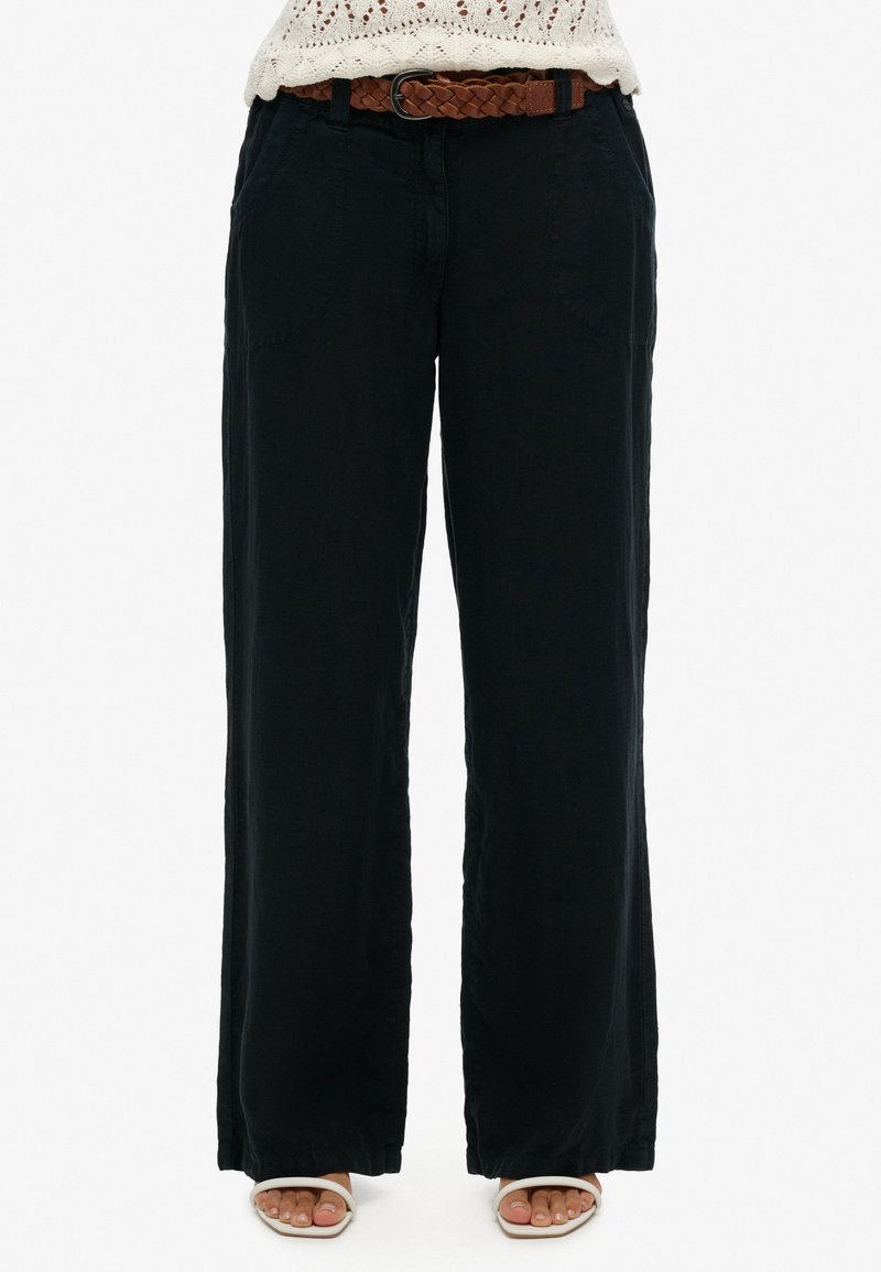 Zwarte, wijde broek van een zachte stof, met twee zijzakken en een gevlochten bruine riem in de taille. Textuur is zichtbaar aan de onderkant.