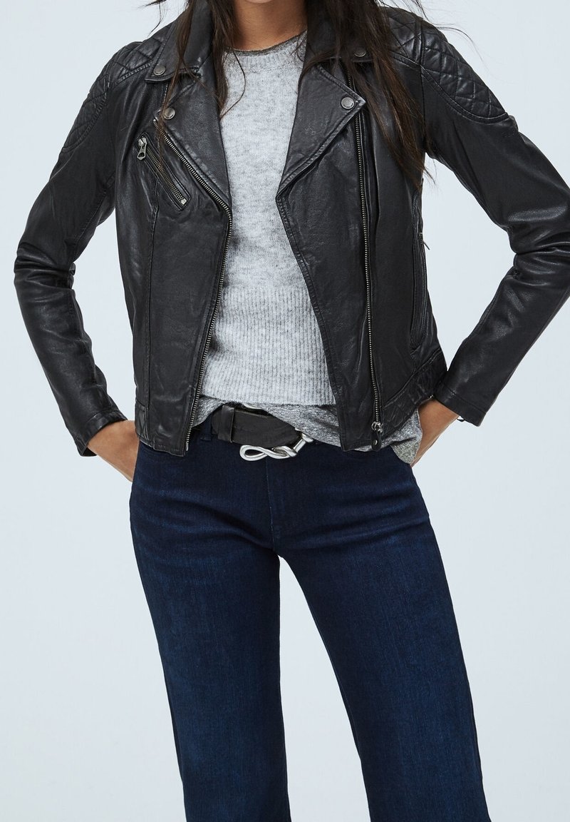 Černá kožená motocyklová bunda s prošívanými ramenními částmi, stříbrnými zipy a slim fit střihem, spárována se šedým svetrem a tmavými džíny.