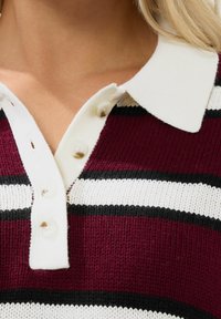 Maroon- och vitrandig stickad polo med vit krage, med knappar och textilstruktur. Nära bild av halsringningen.