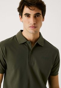 Oliven grøn polo t-shirt med krave, korte ærmer og to knaplukninger. Har diskret branding på brystet. Glat stofstruktur.