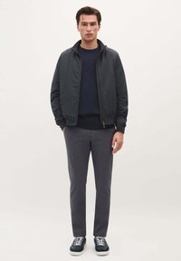 Granatowa kurtka na zamek z wysokim kołnierzem, noszona na granatowym swetrze. Szare spodnie slim fit i ciemne sneakersy z białymi sznurówkami. Gładkie tło.
