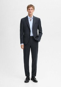 Dark suit with a fitted blazer and matching trousers, paired with a light blue shirt. Glossy black dress shoes complete the look.