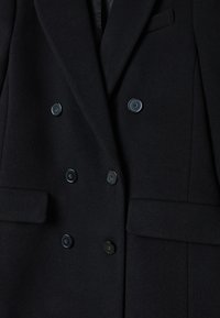 Blazer noir croisé en tissu texturé, doté de six boutons noirs, deux poches avant et d'une coupe ajustée.