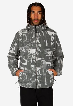 Man wearing a gray patterned hooded jacket with abstract white and black graphic designs, hands in side pockets, facing forward.