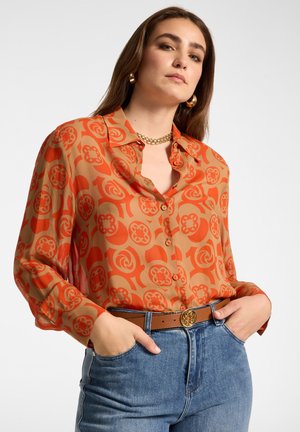 Mujer con blusa abotonada de color naranja con estampado, joyas de oro y vaqueros azules, de pie con las manos en los bolsillos frente a un fondo liso.