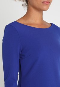 Haut à manches longues bleu avec un col rond, fabriqué en tissu lisse. Présente une silhouette cintrée avec des détails de couture sur les manches.