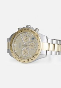 Orologio cronografo in acciaio inox con accenti dorati, quadrante beige, tre sottodial, tachimetro e design del bracciale a maglie.