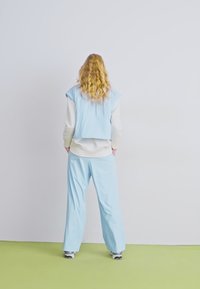 Chaleco corto azul claro sobre una camisa blanca de manga larga, combinado con pantalones de pierna ancha azul claro, con cabello rizado visible desde atrás.