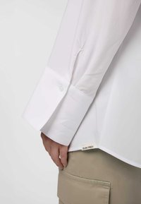 Camicia bianca a maniche lunghe con un ampio polsino, dotata di chiusura con bottone. Etichetta con "SURI FREY" visibile vicino all'orlo, abbinata a pantaloni beige.