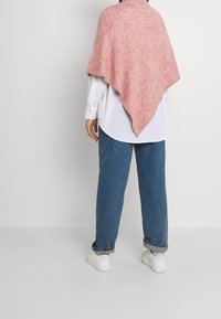 Poncho en maille de mélange rose et blanc, avec une forme triangulaire, une texture douce et un design mi-long, porté sur une chemise blanche et un jean bleu.