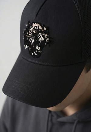 Casquette noire avec une tête de tigre rugissante brodée portée par une personne en sweat à capuche sombre, visage partiellement visible et incliné vers le bas.