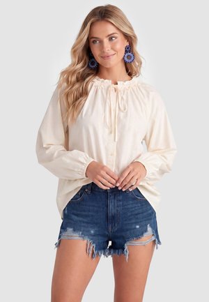 Frau trägt eine cremefarbene Langarmbluse mit Schleife am Hals und blaue, abgenutzte Jeansshorts, ergänzt durch große blaue Ohrringe.