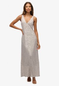 Ärmelloses silbernes Maxikleid mit V-Ausschnitt, das eine schimmernde Paillettenstrukur und einen gestuften Saum aufweist, getragen mit braunen Riemchen-Absatzschuhen.