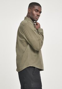Cămașă verde măslinie cu mâneci lungi și guler cu nasturi, având o croială lejeră și detalii vizibile de cusătură. Purtați cu pantaloni cargo negri.