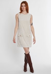 Beiges ärmelloses Kleid aus strukturiertem Stoff mit zwei vorderen Taschen und Akzenten aus Holzknöpfen, kombiniert mit hohen braunen Lederstiefeln.