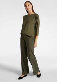 Blusa verde oliva con scollatura a barca, maniche a tre quarti e vestibilità ampia, abbinata a pantaloni dritti coordinati. Indossati con ballerine nere.