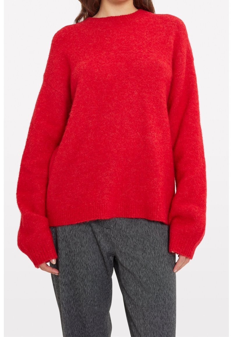 Vrouw in een losse rode fuzzy trui met lange mouwen, gecombineerd met grijze gestructureerde broek, die net boven de schouders eindigt.