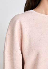 Lichtroze sweatshirt met een gestructureerde afwerking, voorzien van kleine, multikleuren stippen. De brede halslijn en verlaagde schouders verhogen het comfort.