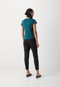 Teal T-skjorte med kort erme og blonder, parret med sorte slim-fit bukser og kraftige hælklip, sett fra baksiden.