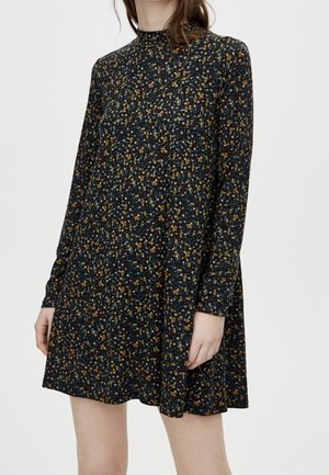 Kvinde iført en sort kjole med lange ærmer og høj hals med et lille flerfarvet blomsterprint, stående mod en ensfarvet baggrund.