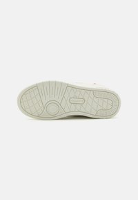 Suole delle scarpe con un design in gomma testurizzata, prevalentemente beige chiaro, dotate di un emblema circolare e un motivo a griglia per una migliore trazione.