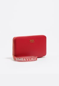 Sarkans ādas maks ar teksturētu virsmu, zelta logotips un atvienojams siksniņš, uz kura kontrastējošā krāsā rakstīts "BIMBA Y LOLA".