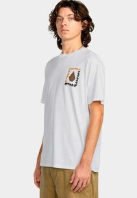Joven con cabello rizado que lleva una camiseta blanca con un pequeño logo de tablero de ajedrez y texto en el lado izquierdo del pecho, combinada con pantalones marrones.