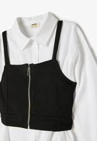 Camiseta corta negra con tirantes en los hombros y un cierre frontal sobre una camisa blanca con cuello. Presenta dos bolsillos frontales en la camiseta.
