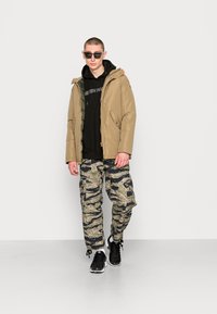 Giacca puffer beige sopra un felpa nera, abbinata a pantaloni cargo camouflage. Il modello indossa scarpe nere e occhiali da sole scuri.