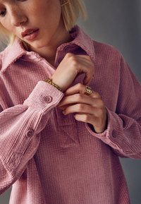 Chemise en velours côtelé violet avec un col boutonné, présentant des boutons sur le devant et des manches texturées. Accents de bijoux dorés comprenant un bracelet et une bague.