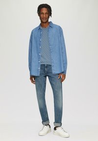 Lichtblauwe denimbloes met lange mouwen, dichtgeknoopt. Grijze trui met stippen eronder, blauwe spijkerbroek en witte sneakers met zwarte accenten.