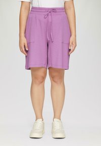 Lila Shorts aus strukturiertem Material, ausgestattet mit einem elastischen Bund mit Kordelzug und zwei vorderen Taschen. Getragen mit weißen Sneakers.