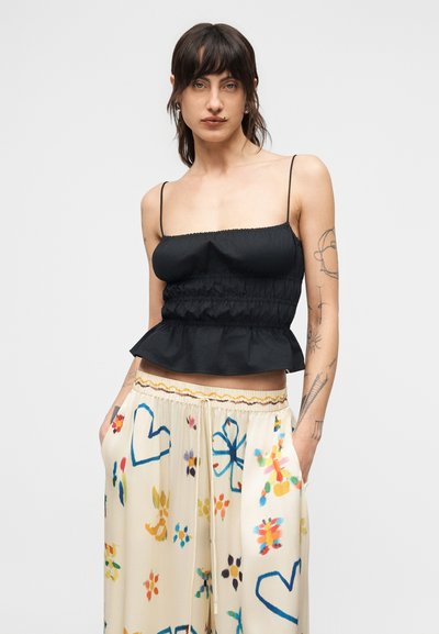 Νεαρή γυναίκα με σκοτεινά μαλλιά που φοράει μαύρο ριχτό αμάνικο τοπ με ρυτιδώσεις και άνετο κρεμ παντελόνι με πολύχρωμα αφηρημένα σχέδια, στέκεται με τα χέρια στις τσέπες.