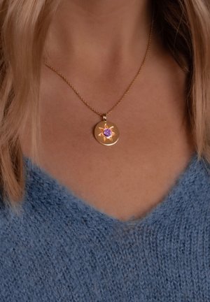 Guldhalskæde med en rund medaljon, der har et stjernes design og en lilla ædelsten i midten, båret over en blå strikbluse.