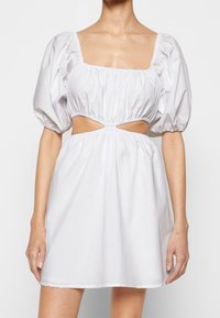 Vit klänning med puffärm och fyrkantig halsringning, elastisk midja och utskärningsdetalj. Tillverkad av glatt tyg med en flödande silhuett.