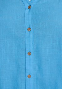 Hellblaues Leinenhemd mit V-Ausschnitt, ausgestattet mit Holzknöpfen entlang der Vorderseite und einem dezenten strukturierten Stofffinish.