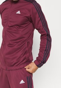 Homme portant un survêtement Adidas bordeaux avec des bandes noires sur les manches et les jambes, main reposant sur la hanche sur un fond uni.
