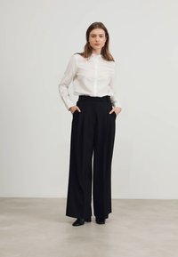 Camicia bianca con un leggero lucido, infilata in pantaloni neri a vita alta e a gamba larga. Scarpe nere; il modello sta in piedi con le mani nelle tasche.