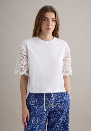 Giovane donna che indossa un top bianco con maniche in pizzo e pantaloni blu con fantasia e coulisse, in piedi davanti a uno sfondo neutro.