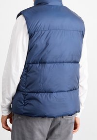 Námořnicky modrá prošívaná vesta se stojáčkem, s prošívanými sekcemi a hladkou texturou, nošená přes bílou košili s dlouhým rukávem.