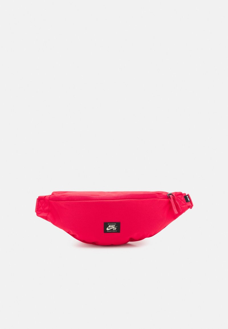 Sac banane rouge Nike SB avec patch logo à l'avant et fermeture éclair supérieure sur fond blanc uni.