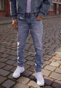 Ljusblå denimjeans med smal passform, framfickor och en subtil blekning, i kombination med vita sportskor på en kullerstensgata.